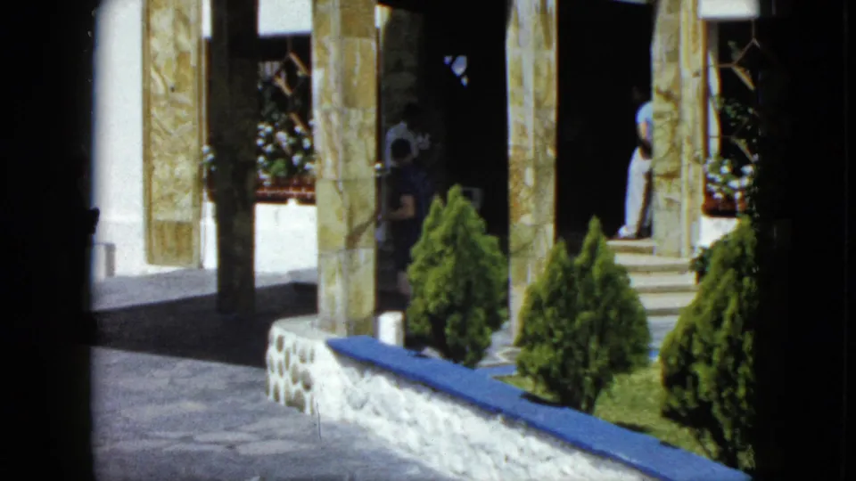 Wide view of the sun-lit courtyard of an upscale building in Acapulco, Mexico, circa 1962, with stone columns, blue-trimmed ledge, topiaries, and figures in background.