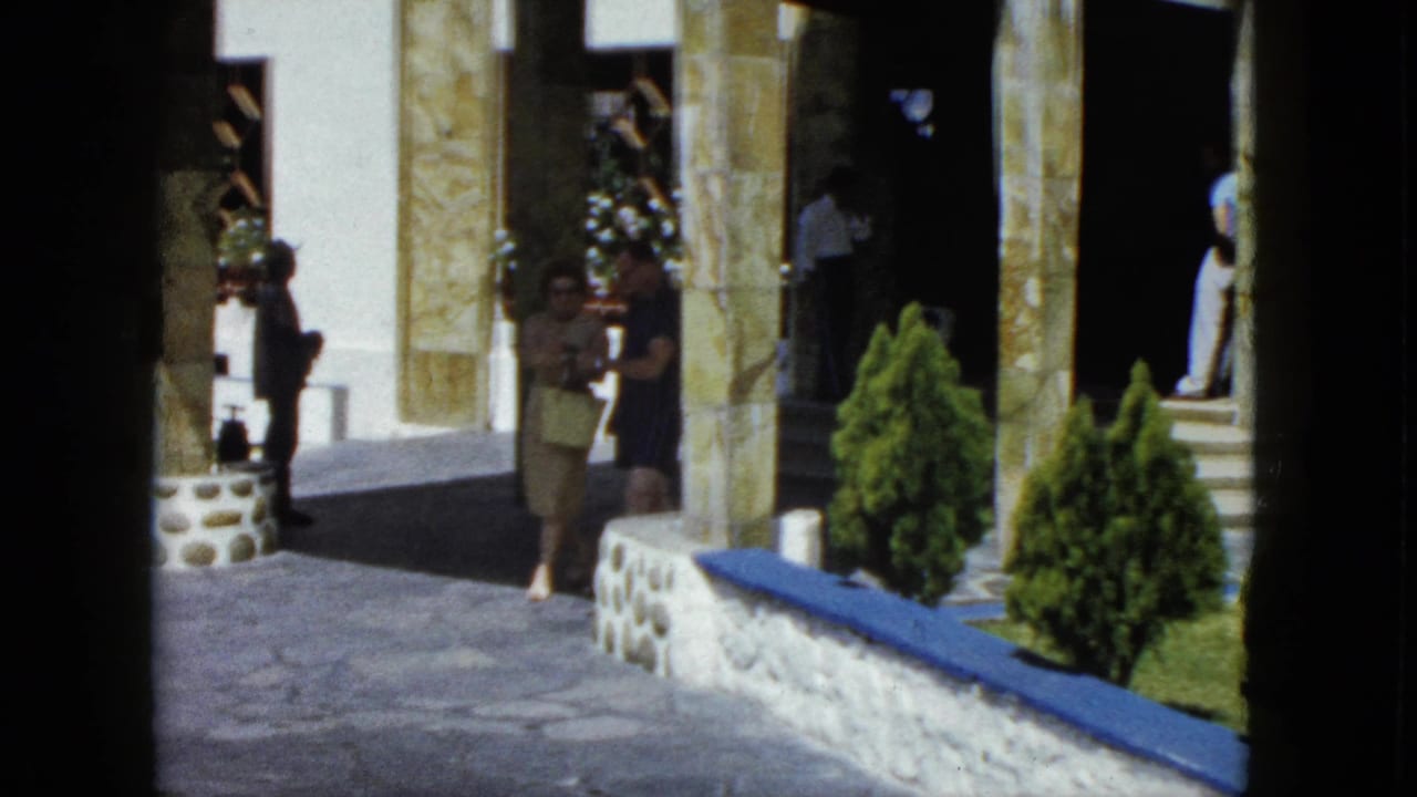 Person walking toward the entrance of an ornate stone-columned building in Acapulco, circa 1962, with conical shrubs and blue ledge visible.
