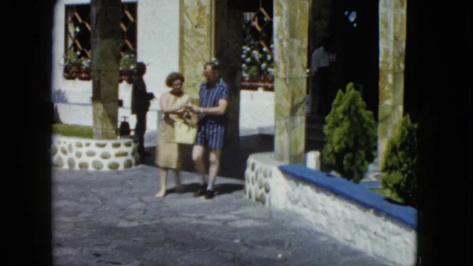 Couple walking toward camera in a resort courtyard in Acapulco, circa 1962; woman in light dress with handbag, man in blue striped shirt and shorts.