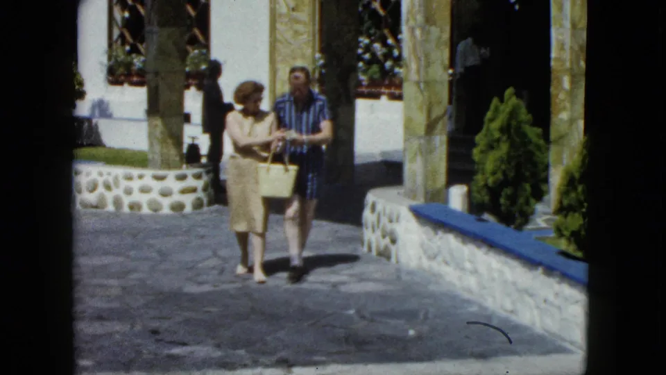 Couple in 1960s leisure dress walking through the courtyard of an upscale building in Acapulco, circa 1962; woman carries a straw or canvas tote.