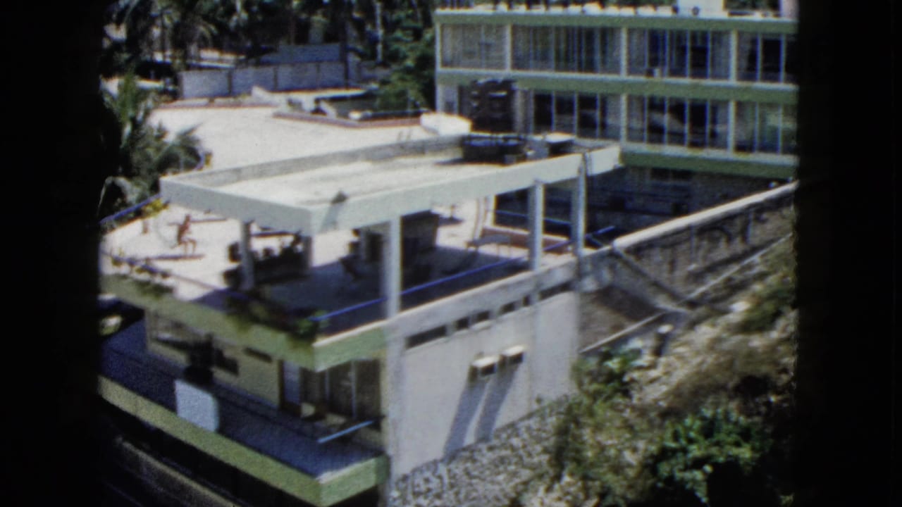 Modernist hillside building with glass windows, flat patio roof, palms, and distant figures.