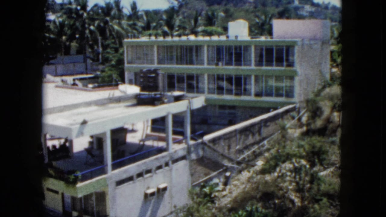 1962 Acapulco frame with a modernist building, wide window bands, flat-roof patio, palms, hillside plants, and tiny figures near the terrace.
