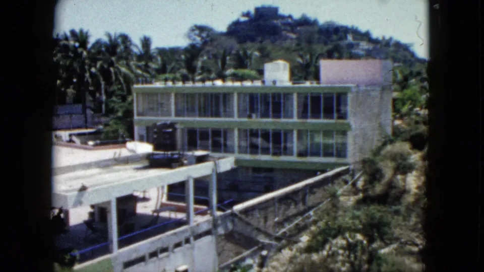 1962 Acapulco home-movie still showing the building's glass facade, patio roof, tropical palms, hillside setting, and terrace edges.