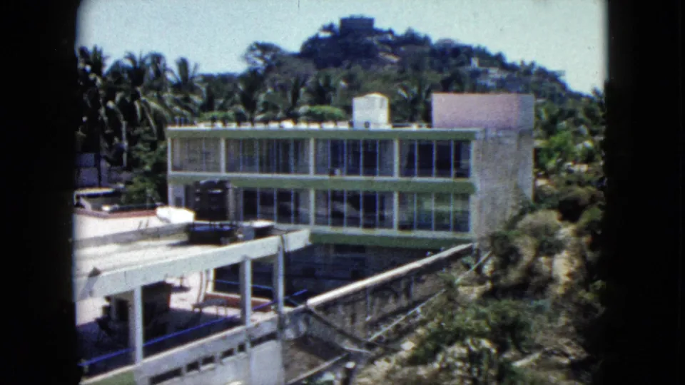 1962 Acapulco frame showing a modernist hillside building with long window rows, a flat patio canopy, palms, and surrounding greenery.