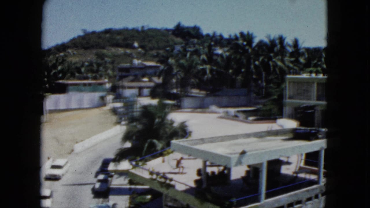 1962 Acapulco home-movie frame showing white flat-roofed buildings, palms, hillside greenery and cars on a sunlit road.