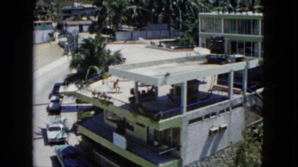 1962 Acapulco home-movie view of a white flat-roofed residence with palms, parked cars and a road below.