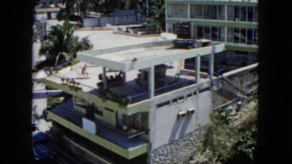 1962 Acapulco frame showing a sunlit modern building, roof terrace area, palms, cars and dense greenery nearby.