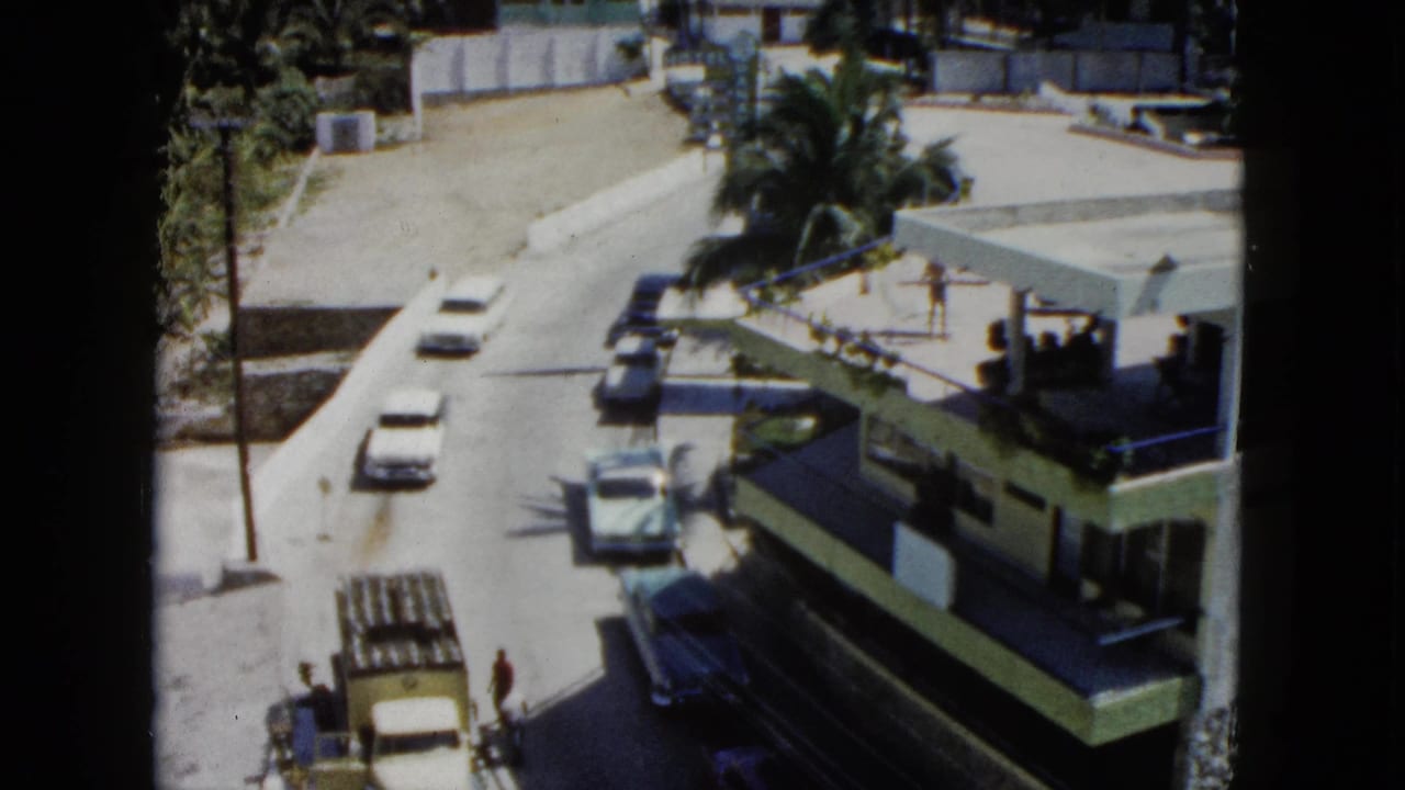 Elevated view of a sunlit street with cars, palms, a low building, and a yellow truck.
