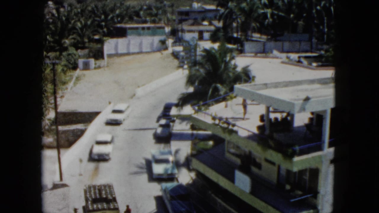 1962 Acapulco street view from above, with several cars on the road, palms, a low building, and hillside vegetation behind it.