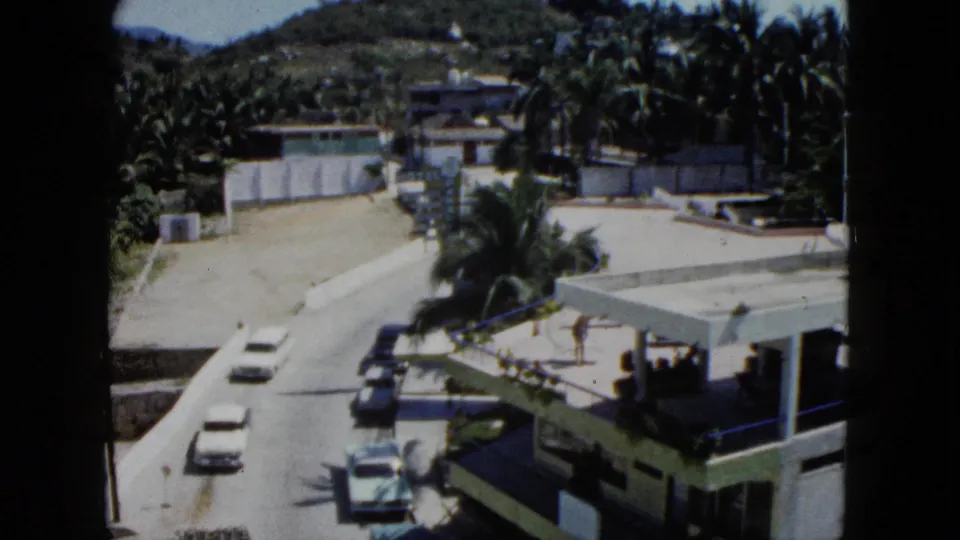1962 Acapulco frame with the road rising through the scene, showing cars, a yellow truck, palms, low buildings, and a wooded hillside.