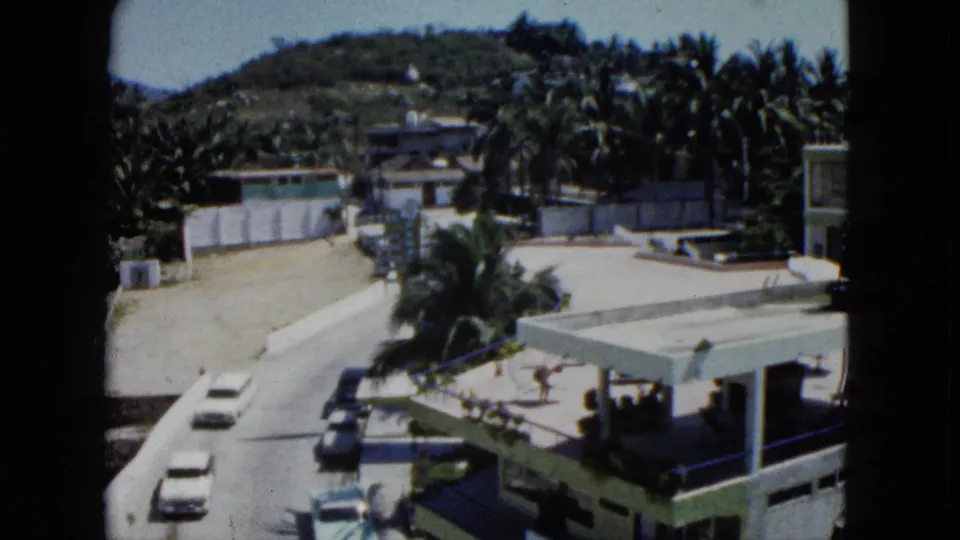1962 Acapulco home-movie view of cars along a sunlit road, with palm trees, terrace railings, low buildings, and green hillside beyond.