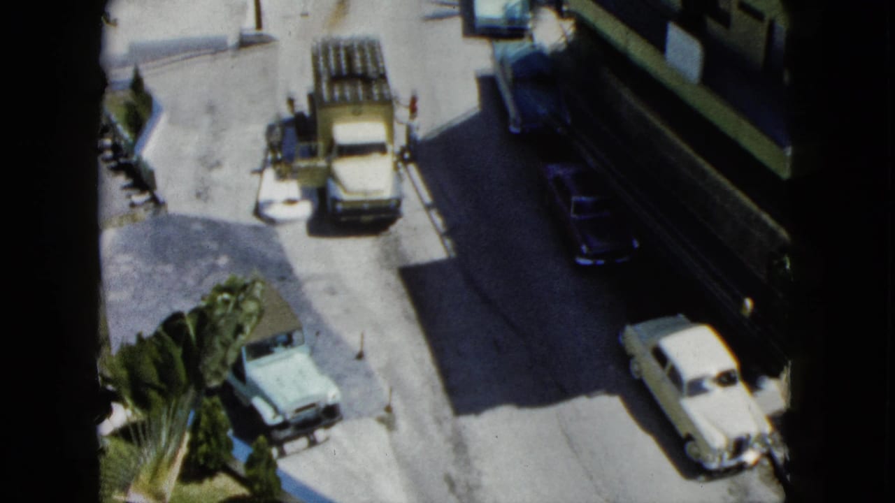 Sunlit street with cars, a truck, pedestrians, palms, and a building balcony.