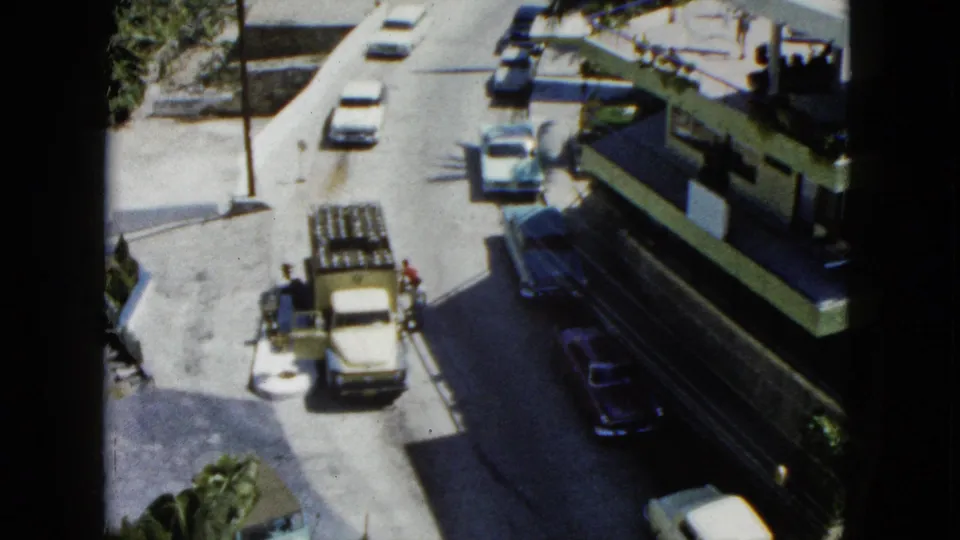 1962 Acapulco frame from above showing a street corner with several cars, a utility truck, pedestrians, palms, and deep midday shadows.