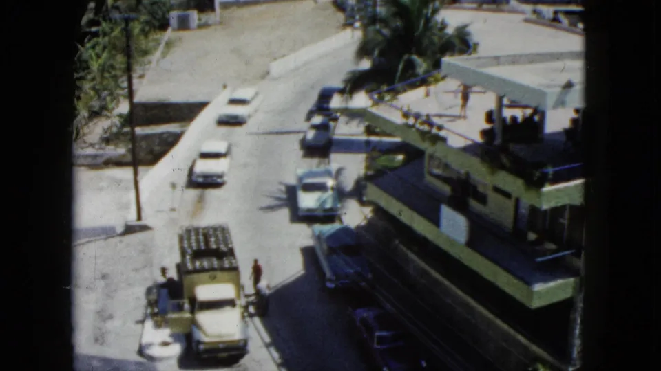 1962 Acapulco home-movie frame showing cars and pedestrians near a flat-roofed building, palm trees, and bright street pavement.
