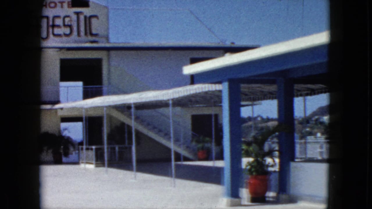 1962 Acapulco frame with the Hotel Majestic sign partly visible above a covered walkway, blue columns, stairs, potted plants, and hillside beyond.
