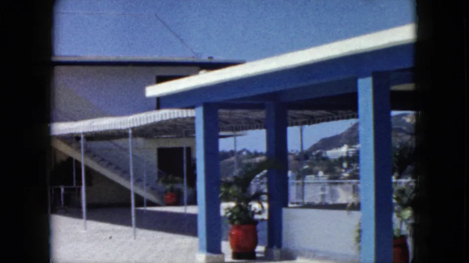 1962 Acapulco home-movie view across the hotel walkway, showing blue structural columns, white walls, potted plants, railing, and hills in the distance.