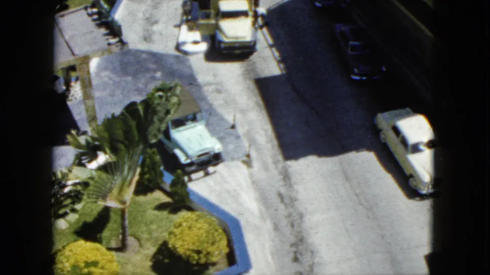 1962 Acapulco frame looking down from the hotel toward a street with classic cars, curbside parking, palm landscaping, shrubs, and bright pavement.