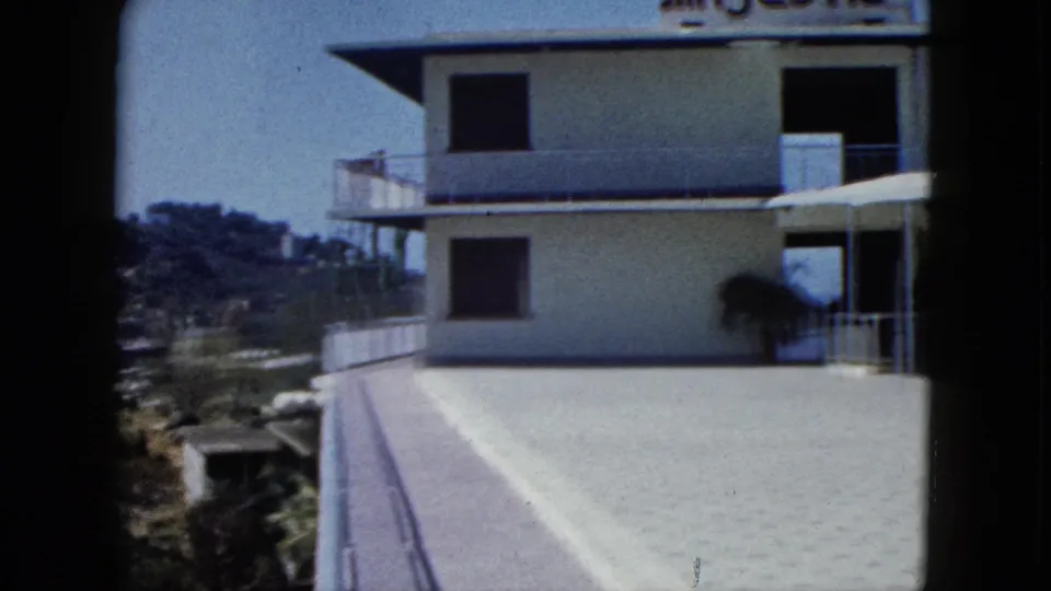 1962 Acapulco frame with a sunlit terrace, white building facade, dark window openings, and hillside visible at the left edge.