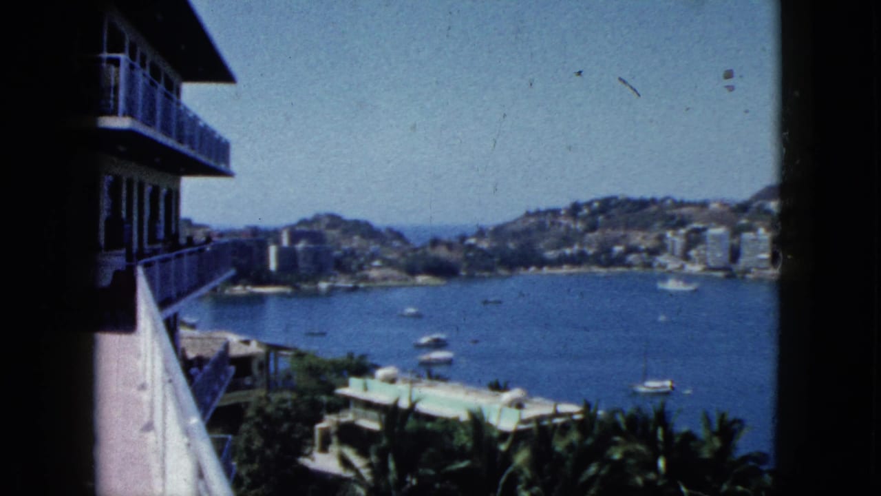 Acapulco Bay seen from a high balcony in 1962, with small boats on the water and buildings along the hillside shore.