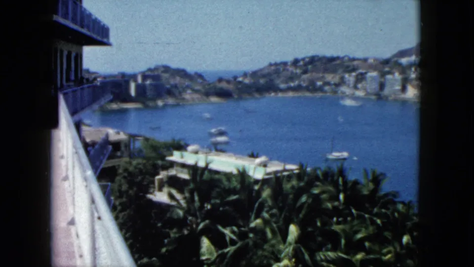 1962 Acapulco home-movie frame showing balcony rail, tropical foliage, bay water, boats, and hillside buildings beyond.