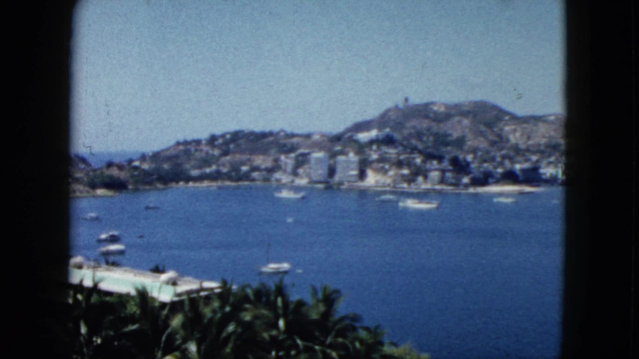 Blue bay with small boats, hillside buildings, palms, and dark balcony or window edges.