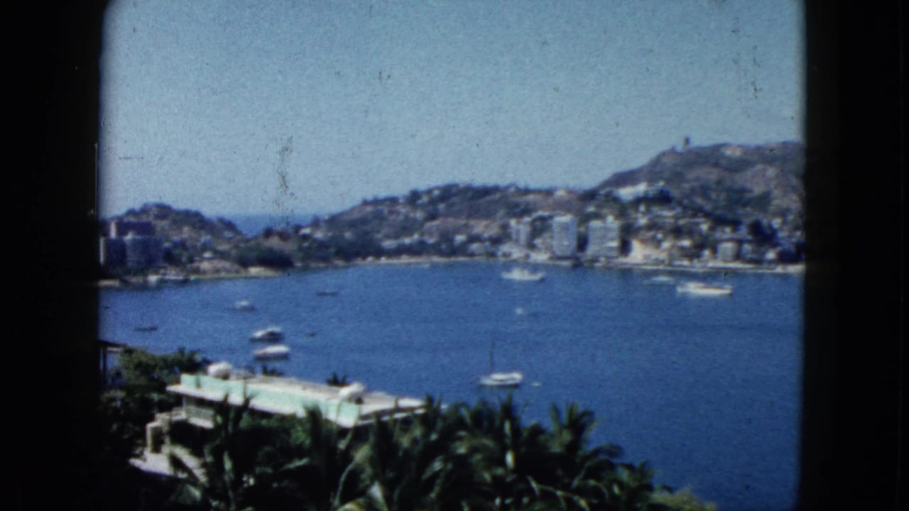 Bay view with anchored boats, waterfront buildings, green palms, hills, and dark vertical frame edges.
