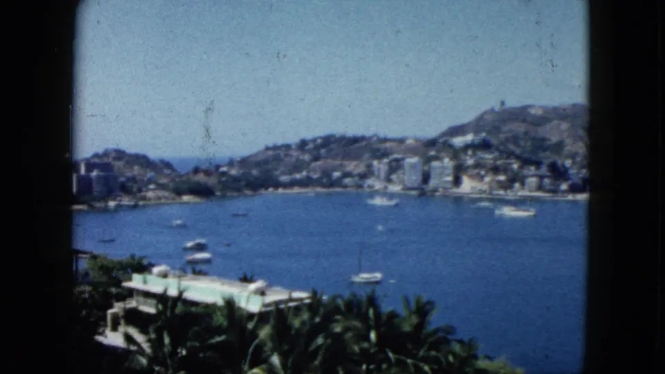 1962 Acapulco bay scene seen from a high balcony or window, with boats on the water and hillside buildings beyond.