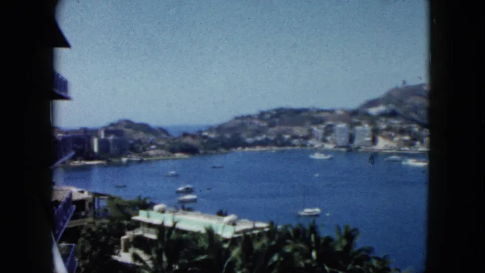 1962 Acapulco home-movie frame showing the bay, boats, palms, a nearby building edge, and hills lined with structures.