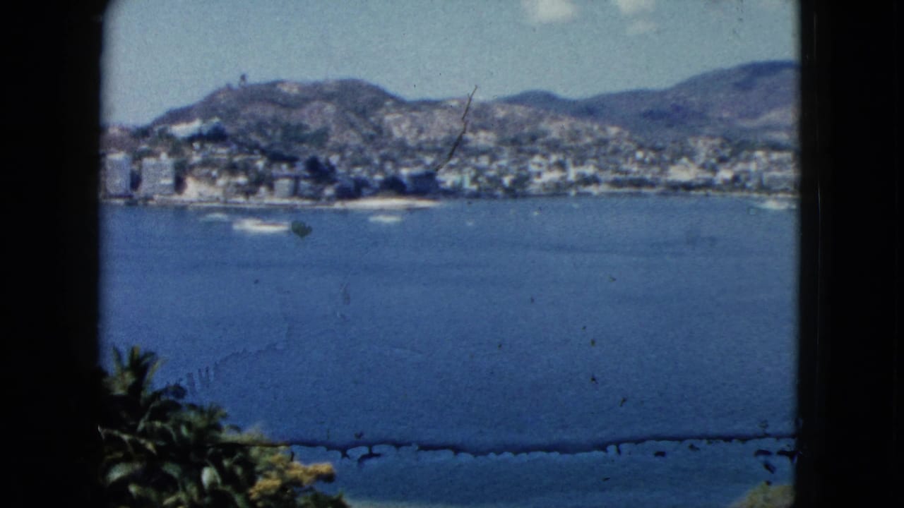 1962 Acapulco bay view with small boats on blue water, hillside buildings, mountains, palms, and dark window edges.