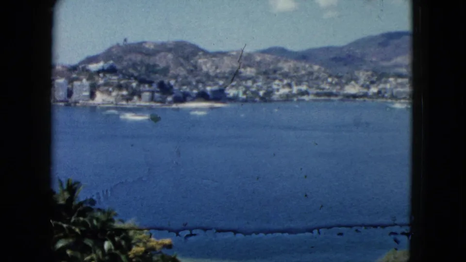 1962 Acapulco bay view with small boats on blue water, hillside buildings, mountains, palms, and dark window edges.