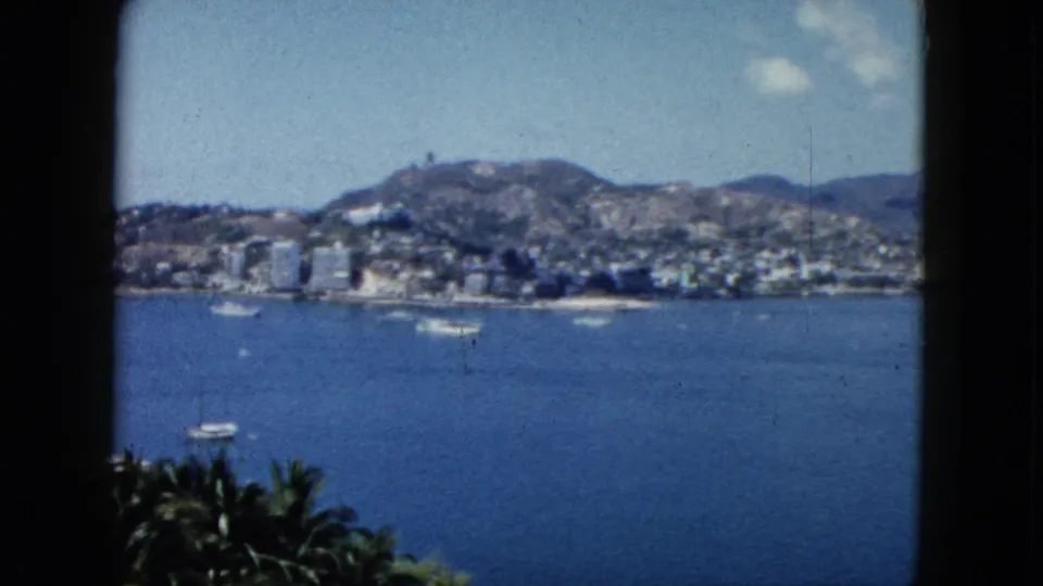 1962 Acapulco bay panorama showing sailboats on blue water, palms in the foreground, and city buildings below mountains.