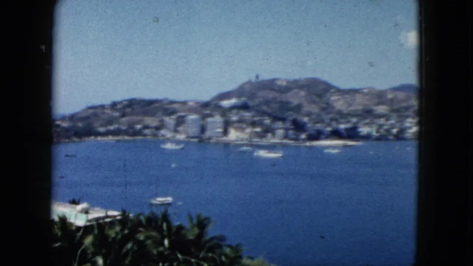 1962 Acapulco view from an elevated shaded vantage point, with boats on the bay, palms below, and mountains behind the city.