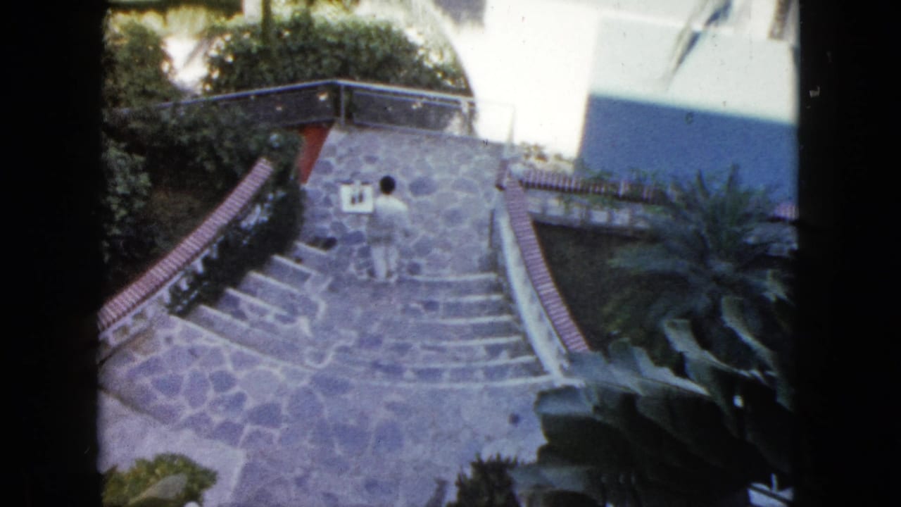In 1962 Acapulco home-movie footage, a man in light clothing walks down stone steps between curved brick railings, shrubs, and palms.