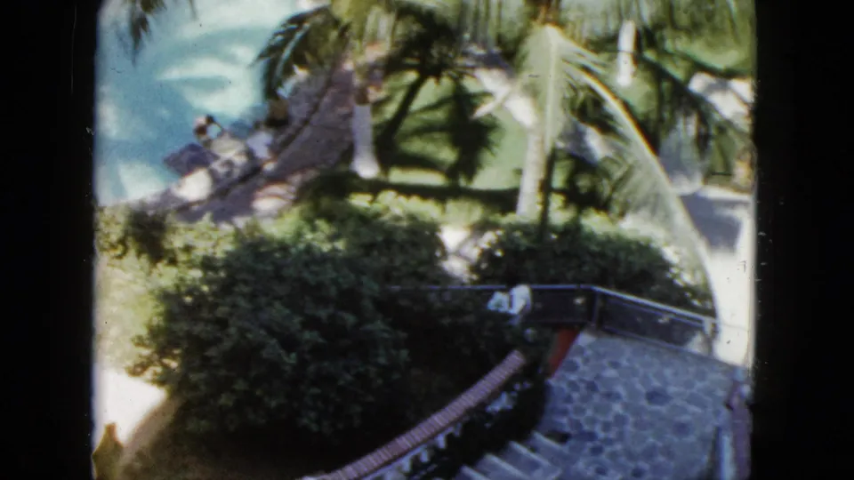 Handheld 8mm footage in Acapulco tilts across palm fronds, dense shrubs, a stone stairway, and a curved railing in a landscaped outdoor setting.