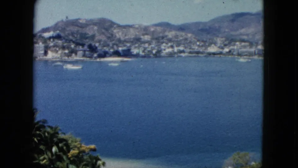 The clip resolves to a 1962 Acapulco bay view, with blue Pacific water, small boats, a hillside coastal town, mountains, and tropical foliage in the foreground.