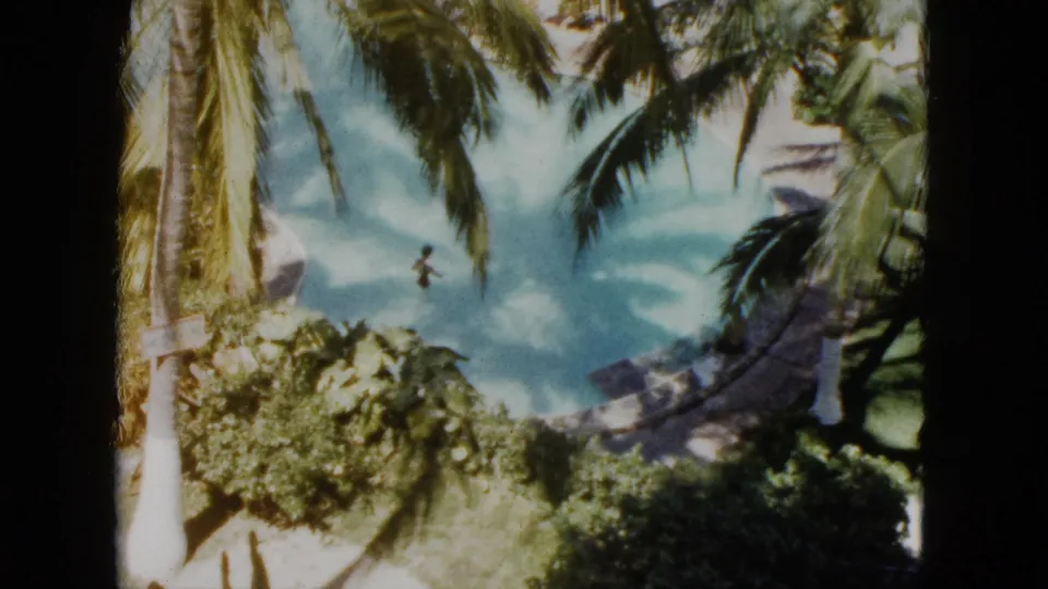 1962 Acapulco resort footage shows a small figure swimming in a blue lagoon below overhanging palms and tropical foliage.