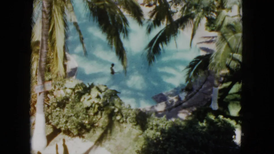 A 1962 Acapulco home-movie view looks down through palm trees toward a swimmer in the resort lagoon and surrounding vegetation.