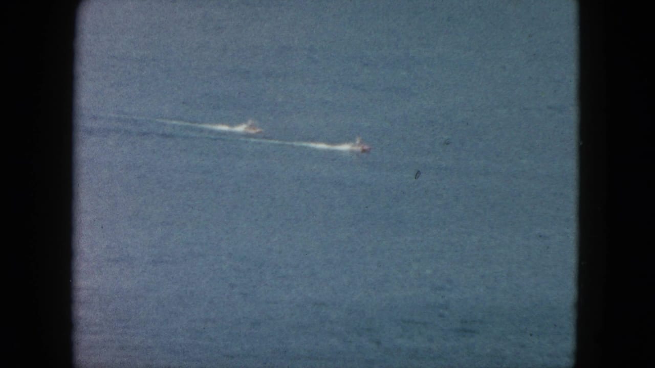 1962 Acapulco footage frame with two distant speedboats crossing blue ocean, each leaving a bright wake line.