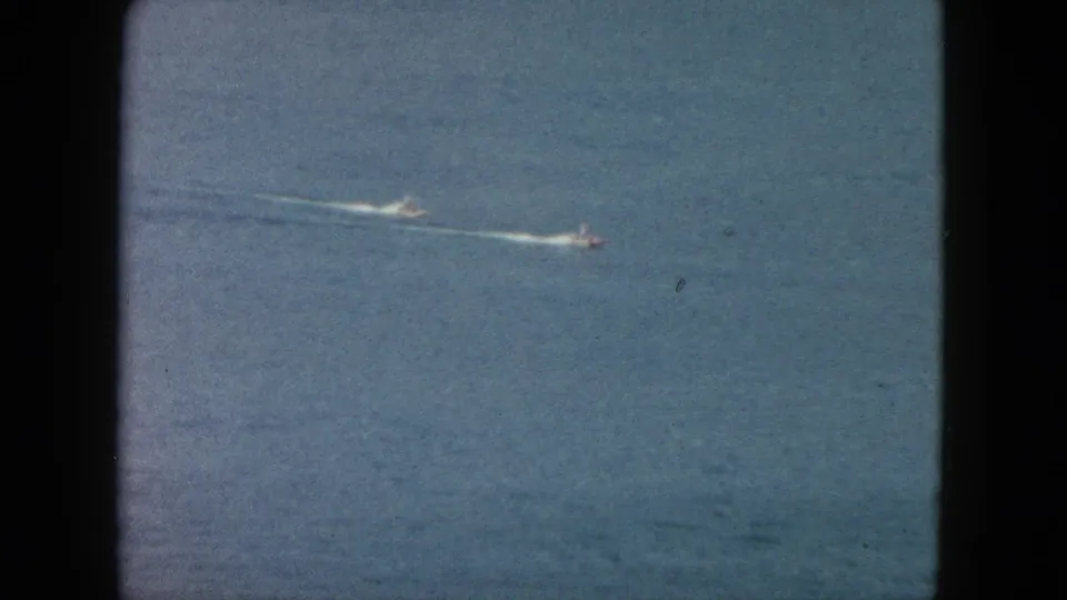 1962 Acapulco footage frame with two distant speedboats crossing blue ocean, each leaving a bright wake line.