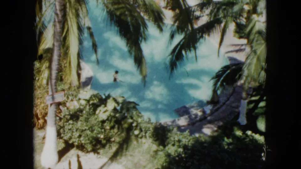 1962 Acapulco home-movie frame showing a palm-lined resort pool with a distant swimmer and surrounding tropical greenery.
