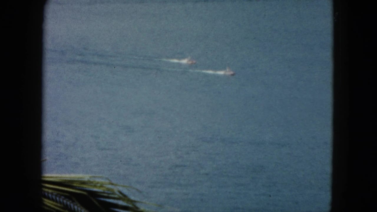 1962 Acapulco 8mm frame showing two distant speedboats moving across blue water with palm fronds in the foreground.