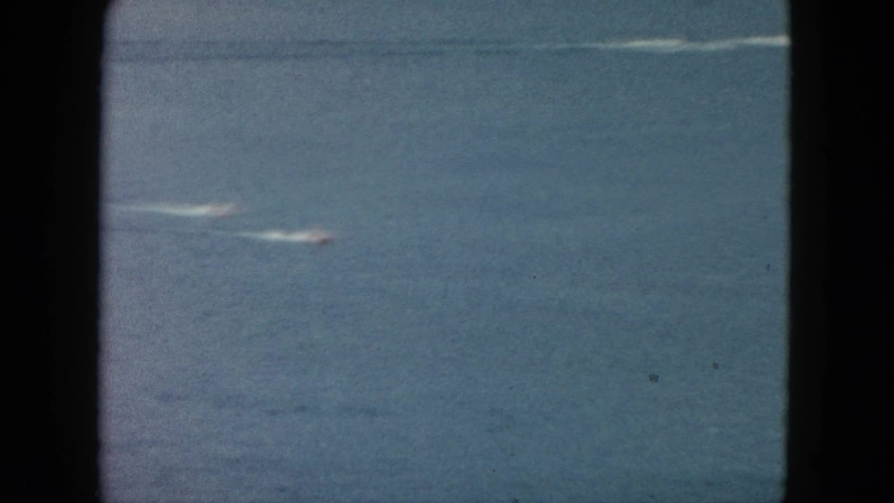 Two distant speedboats streak across open water, leaving pale wakes in a grainy 8mm frame.