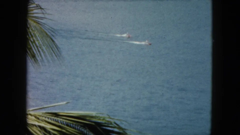 1962 Acapulco home-movie frame with two distant motorboats on turquoise water and palm fronds in the foreground.