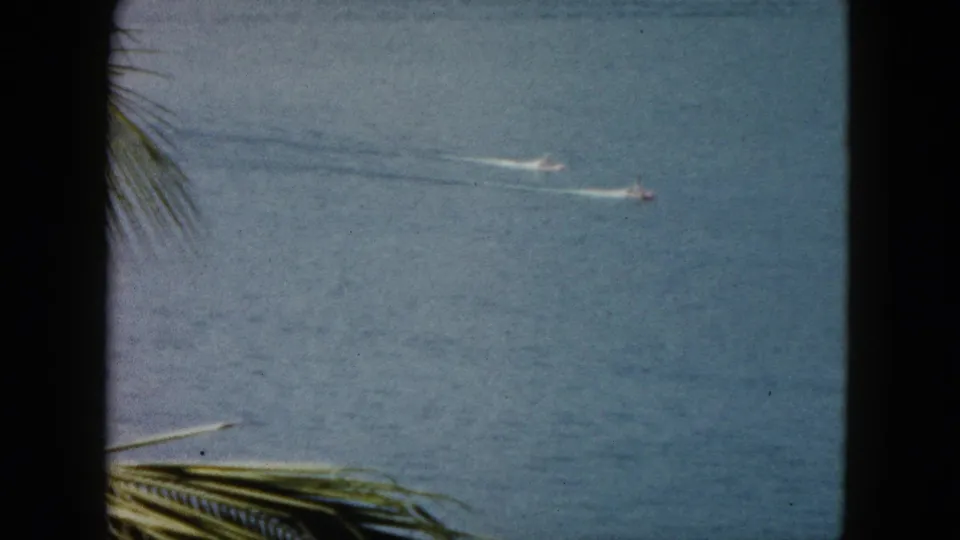 1962 Acapulco home-movie frame showing two small motorboats crossing blue ocean water with palm fronds in front.