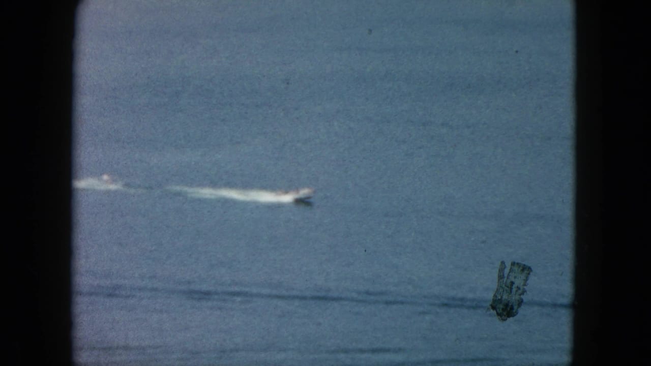 1962 Acapulco home-movie frame showing a small white speedboat moving across blue ocean water behind dark vertical framing.