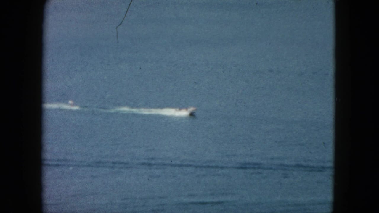 1962 Acapulco 8mm frame with a distant white speedboat racing across open blue water and leaving a long white wake.