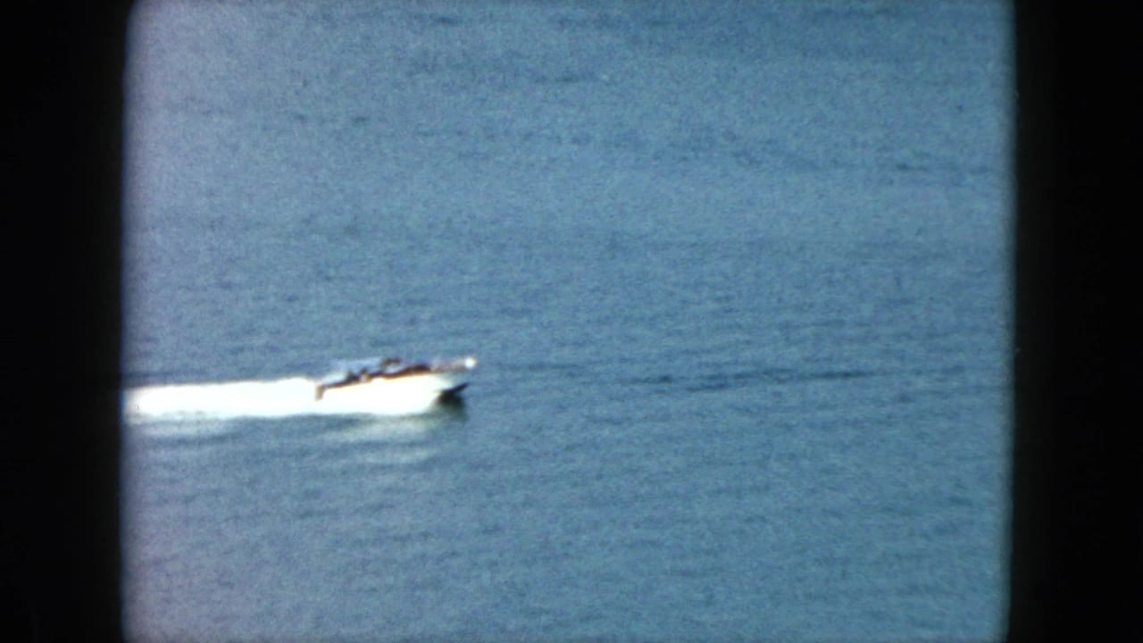 1962 Acapulco home-movie frame of a small white speedboat crossing calm blue water with passengers faintly visible.