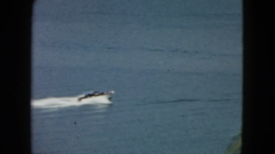 A white speedboat passes low in the 8mm frame on blue Acapulco water, with passengers indistinct and a bright wake behind.