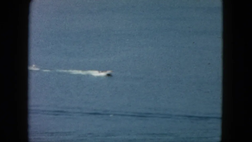 Distant 1962 Acapulco speedboat receding across open water, leaving a pale wake in a grainy 8mm frame.
