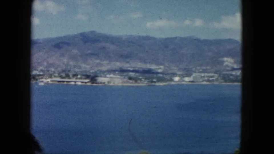 1962 Acapulco home-movie view of blue bay water with a distant shoreline and mountains beyond the dark film-frame edge.